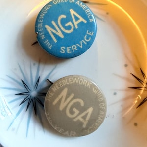 May include: Two circular pinback buttons featuring the text The Needlework Guild of America. One button is blue with white text reading NGA Service. The second button is grey with white text reading NGA and 2nd Year. Both have a pin backing.