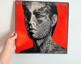 May include: A framed album cover for The Rolling Stones' "Tattoo You." The artwork features a black and white portrait of a person with intricate tattoo designs on their face and neck, set against a vibrant red background.