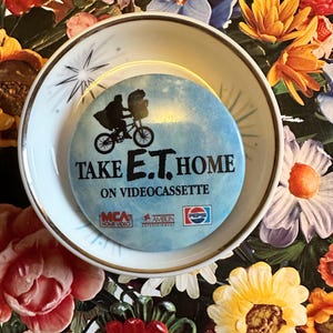 May include: A round, light blue button with the text "TAKE E.T. HOME ON VIDEOCASSETTE" and a silhouette of E.T. on a bicycle. The button is on a white plate with a gold rim, set against a floral background.