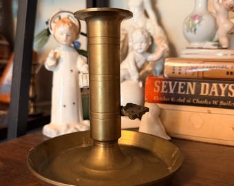 May include: A brass chamberstick candle holder featuring a wide circular drip tray base, a cylindrical stem with horizontal ring detailing, and a small decorative thumb piece attached to the side for carrying the object by hand.