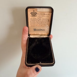 May include: An open, vintage black watch box with gold trim, held by a hand. The box contains a black velvet interior and a printed certificate from Hamilton Watch Co. The certificate includes text about the watch movement.