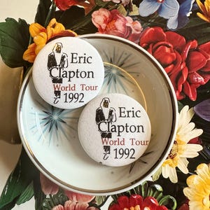 May include: Two white Eric Clapton World Tour 1992 buttons rest in a decorative bowl with a gold rim. The buttons feature a black and white image of Eric Clapton. The bowl sits on a floral background with red, yellow, and blue flowers.