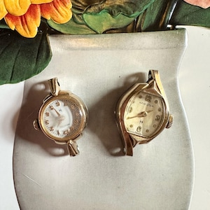 May include: Two vintage gold-toned Hamilton wristwatches. One watch has a white face with black numerals and a gold-toned bezel. The other watch has a cream face with black numerals and a gold-toned bezel. Both watches have a gold-toned crown.