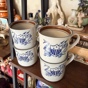 May include: Four white ceramic mugs with a blue and brown design. Each mug features a blue emblem with stars and stripes, and a brown rim. The mugs are stacked on a wooden shelf.