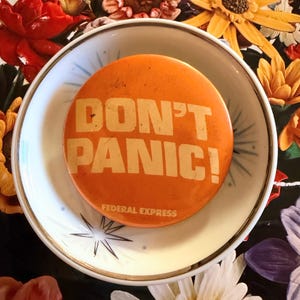 May include: An orange button with the words "DON'T PANIC!" in white block letters. The button also has the words "FEDERAL EXPRESS" printed on it. The button is in a white bowl with a starburst design, surrounded by colorful flowers.