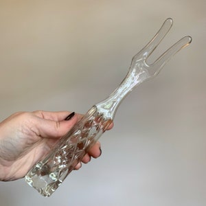 May include: Clear glass serving fork with a diamond pattern on the handle. The fork has two tines and is held in a hand. The glass has a slight iridescent sheen. The serving utensil is likely for a salad or other dish.