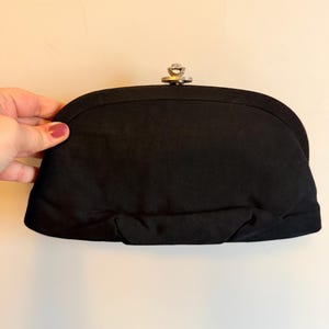 May include: Black clutch with a silver clasp. The clutch is a rounded rectangular shape and appears to be made of a soft fabric. The clasp is a silver-colored metal with a decorative design. The clutch is held by a hand with painted nails.