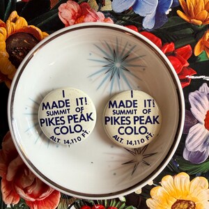 May include: Two vintage white pinback buttons with the text "I MADE IT! SUMMIT OF PIKES PEAK COLO. ALT. 14,110 FT." are displayed inside a white bowl with a gold rim. The background is a floral pattern.