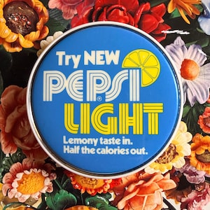 May include: A round, blue Pepsi Light sign with white and yellow text, including a lemon graphic. The sign is surrounded by a variety of colorful, artificial flowers in shades of red, orange, yellow, and purple.