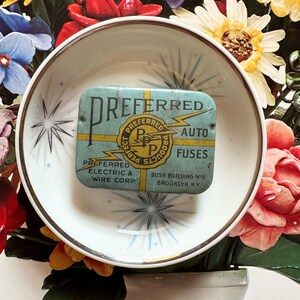 May include: A vintage, rectangular tin of "Preferred Auto Fuses" in a light blue hue, resting in a white and gold-rimmed bowl. The tin features gold and black text and a lightning bolt graphic. The bowl is set against a backdrop of colorful flowers.