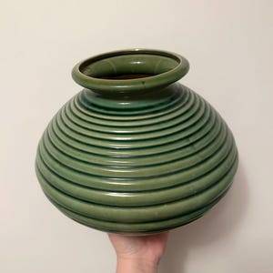 May include: A green ceramic vase with a bulbous body and a narrow, flared opening. The vase features a series of horizontal, ridged lines that encircle the body. The vase is held by a hand against a plain white background.