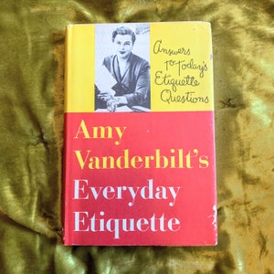May include: A red and yellow book titled "Amy Vanderbilt's Everyday Etiquette" with a photo of a woman on the cover. The text "Answers to Today's Etiquette Questions" is written in cursive on the cover.