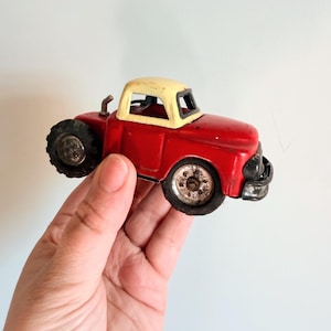 May include: A vintage red toy truck with a cream-colored cab. The truck has black tires and a small exhaust pipe. The toy truck is a classic design, likely from the mid-20th century, and shows signs of wear.