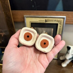 May include: Two ceramic salt and pepper shakers with brown rubber stoppers on the bottom. The shakers are white with gold accents.