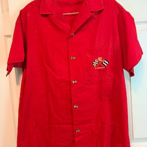 May include: A bright red short-sleeved button-down shirt with a camp collar. The left chest pocket features an embroidered design with the number 40, two crossed flags, and the text R. CABRERA. The shirt has a front button closure.