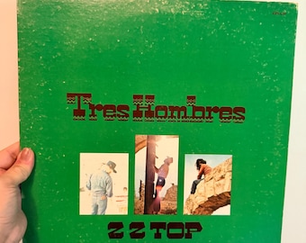 May include: A square vinyl record jacket with a bright green background. The text Tres Hombres is printed in dark brown, followed by three rectangular photographs of people in outdoor settings. Below, the text ZZ TOP and LONDON are visible.