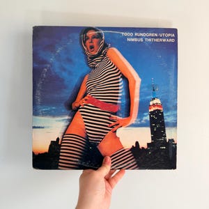 May include: Vintage vinyl record album cover featuring a woman in a striped outfit with a red belt, set against a cityscape and a blue sky. The album title is "Todd Rundgren/Utopia Nimbus Titherward."