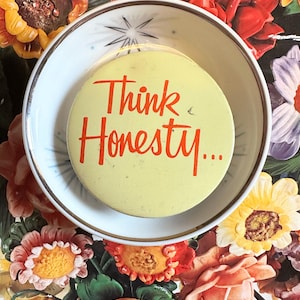 May include: A yellow button with the words "Think Honesty..." in orange script sits inside a white and gray patterned bowl. The bowl is surrounded by a colorful floral arrangement with red, yellow, and pink flowers.