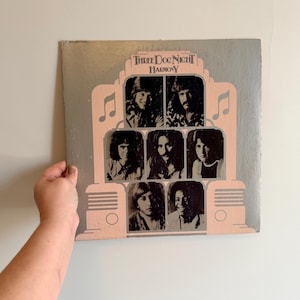May include: Vintage vinyl record album cover for Three Dog Night's "Harmony." The album cover features a light pink and gray design with the band's name and album title. The cover has a retro design with images of the band members.