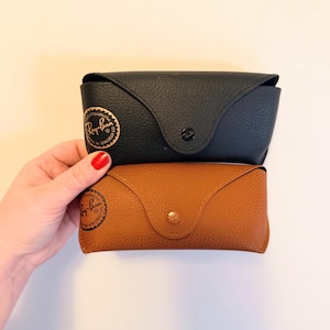 May include: Two Ray-Ban sunglasses cases, one black and one brown, are stacked. The black case has a gold logo and a snap closure. The brown case has a gold snap closure and a similar logo. Both cases are made of a textured material.