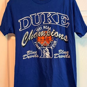 May include: Royal blue t-shirt featuring the text "DUKE Champions 1991 NCAA Final Four" in white and gold. The shirt also displays the "Blue Devils" logo and text. A classic sports apparel item.