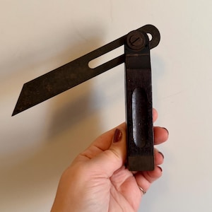 May include: A vintage adjustable bevel with a dark wooden handle and a dark metal blade. The blade is angled, and the tool is held in a hand against a neutral background. This woodworking tool is used for measuring and marking angles.