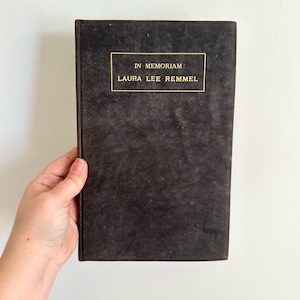 May include: A dark brown book with the title "IN MEMORIAM" and the name "LAURA LEE REMMEL" in gold lettering. The book has a textured cover and is held by a hand.