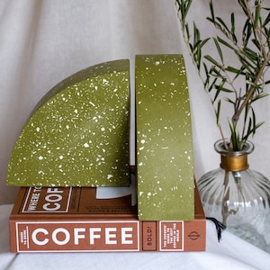 May include: Olive green bookends with white speckles, resting on a stack of books with the word "COFFEE" visible. A glass vase with a gold rim and a sprig of greenery is in the background.