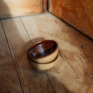 May include: Two small, round ceramic bowls stacked on top of each other. The bowls feature a smooth, dark brown glazed interior and a textured, light tan matte exterior finish. The design is simple and functional, showcasing a contrast between the glossy inner surface and the earthy, natural tone of the outer clay body.