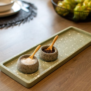 May include: A rectangular, sage green serving tray with a speckled pattern holds two small, textured stone bowls. One bowl contains salt, the other peppercorns. Small wooden spoons rest in each bowl. The tray sits on a wooden surface.