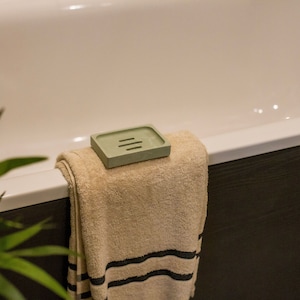 May include: A sage green soap dish with three slots rests on a folded beige towel with black stripes. The towel is on the edge of a white bathtub. The bathroom setting includes a dark brown wall and a green plant.