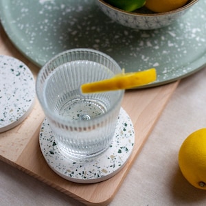 May include: A glass of water with a lemon slice rests on a terrazzo coaster. The coaster has a white base with green flecks. A matching tray and coaster are also present. A whole lemon and a bowl of citrus fruits complete the scene.