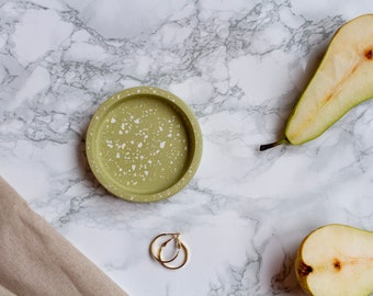May include: A round, olive green trinket tray with white speckles, gold hoop earrings, and two pear halves on a marble surface. A beige linen cloth is in the lower left corner. The tray is likely for jewelry or small items.