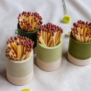 May include: Four small, cylindrical match holders in shades of green and beige, each filled with matches. The match heads are a reddish-brown colour. The holders are arranged on a white surface, with a few daisies scattered around.