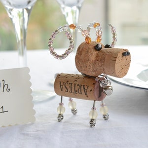 May include: A small, whimsical dog figurine made from a wine cork, beads, and wire. The cork has the word "ARTESA" printed on it. The dog has pink and clear beads for ears and legs.