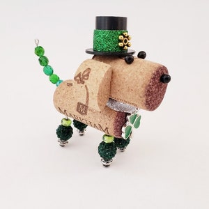 May include: A cork dog figurine wearing a green and black top hat with gold glitter. The dog has a green shamrock on its chest and a silver collar. The dog's tail is made of green beads.