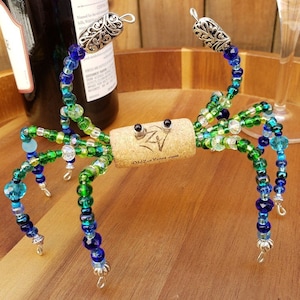 May include: A beaded wine bottle stopper shaped like a crab with green, blue, and clear beads. The crab has silver accents and is sitting on a wooden surface.