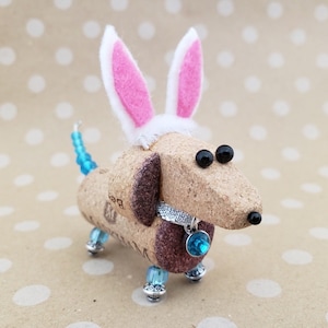 May include: A small, brown dachshund figurine made from wine corks with blue and silver accents. The dachshund is wearing pink bunny ears and a blue gem necklace.