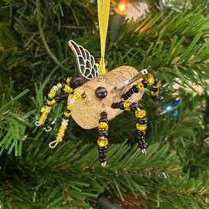 Puede incluir: Un adorno de abeja de corcho con cuentas negras y amarillas, alas plateadas y una antena de alambre. El adorno est&aacute; colgado de un &aacute;rbol de Navidad verde.