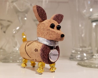 May include: A small cork dog figurine with brown ears, black eyes, and a silver collar with a tag that says "TIMMY". The dog has gold beads for legs and a gold bead tail.