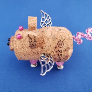 May include: A whimsical pig figurine made from a cork with pink beads for a tail, silver wings, and pink glass eyes. The cork has the number "1" and the text "25-16" printed on it.