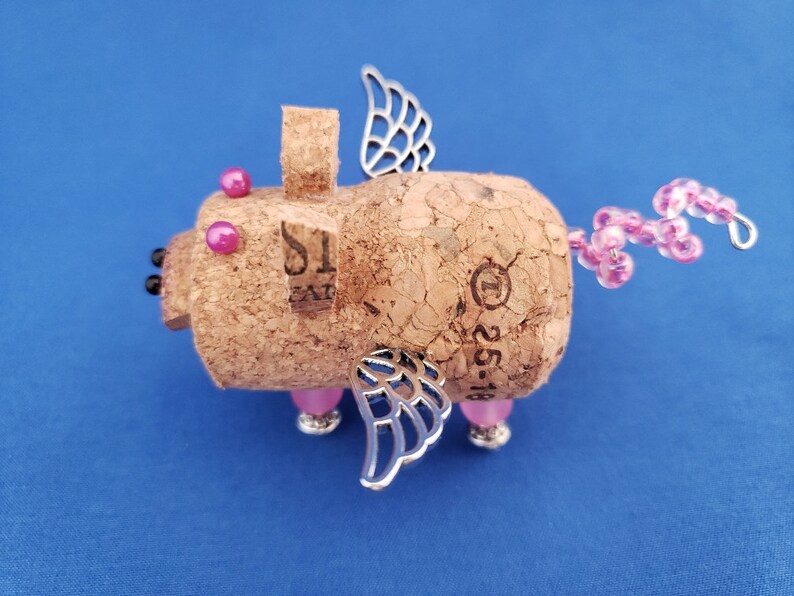 May include: A whimsical pig figurine made from a cork with pink beads for a tail, silver wings, and pink glass eyes. The cork has the number "1" and the text "25-16" printed on it.