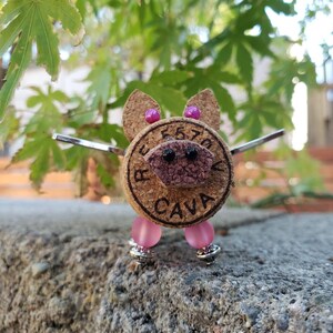 May include: A small pig figurine made from a cork with pink beads for legs and a black nose and eyes. The cork has the words "Reserva Cava" printed on it.