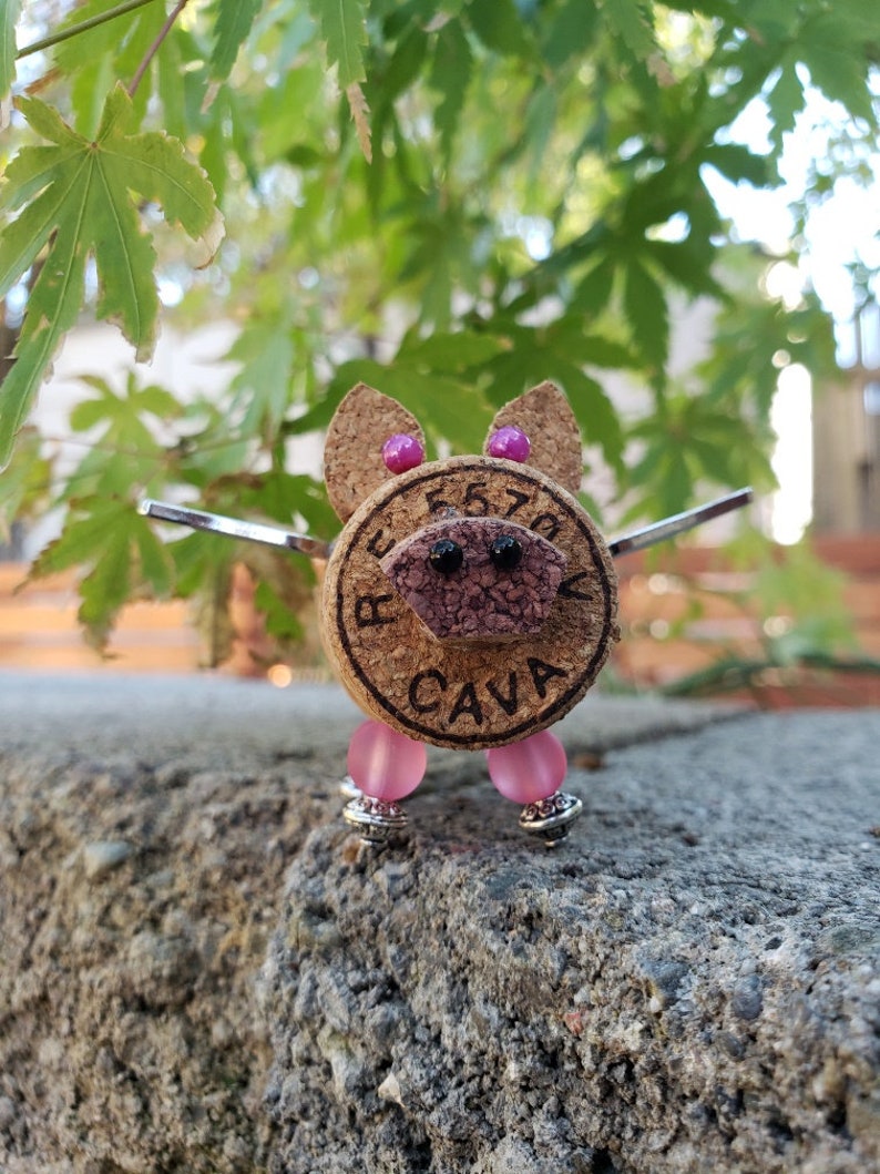 May include: A small pig figurine made from a cork with pink beads for legs and a black nose and eyes. The cork has the words "Reserva Cava" printed on it.