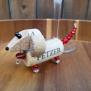 May include: A dachshund-shaped wine charm made from a wine cork with the name "FETZER" written on it. The charm has red beads for a tail and black beads for eyes.