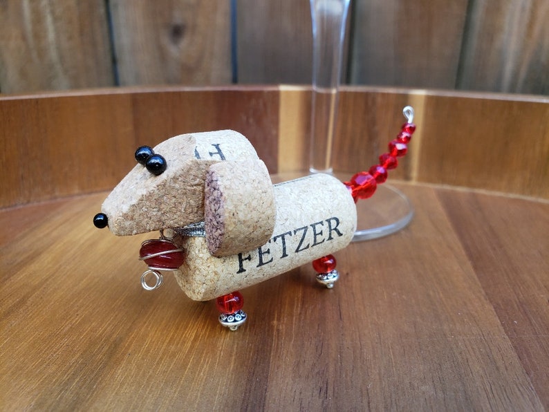 May include: A dachshund-shaped wine charm made from a wine cork with the name "FETZER" written on it. The charm has red beads for a tail and black beads for eyes.
