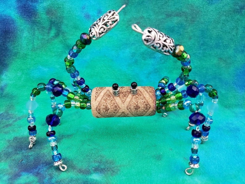 May include: A beaded crab sculpture with a cork body, blue and green glass beads, and silver wire legs. The crab is posed with its claws raised.