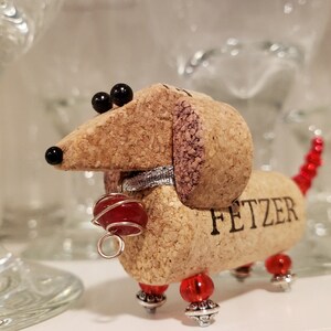May include: A cork dachshund figurine with red beads for a tail and legs. The figurine has black eyes and a silver collar. The cork is engraved with the word "FETZER".