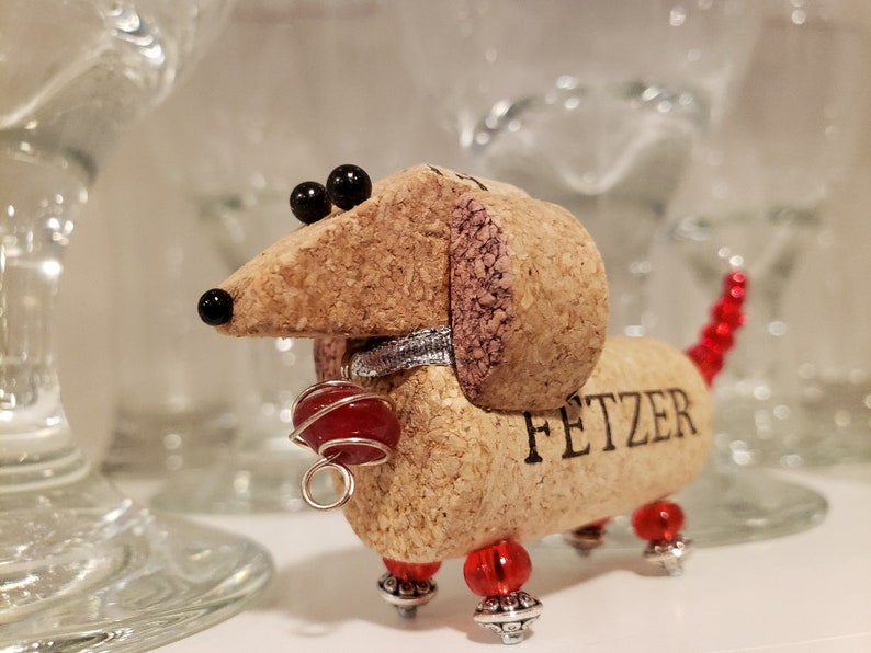 May include: A cork dachshund figurine with red beads for a tail and legs. The figurine has black eyes and a silver collar. The cork is engraved with the word "FETZER".