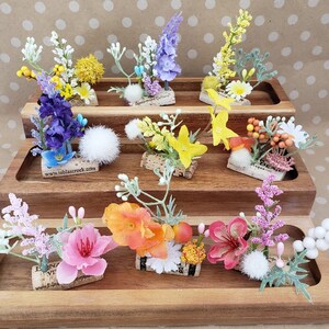 May include: Assortment of small floral arrangements on cork rounds, displayed on a tiered wooden stand. The arrangements feature a variety of flowers, including purple, yellow, pink, and orange.